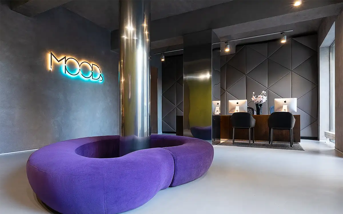 Moderne Rezeption im MOODs Boutique Hotel Prag mit violettem Rundsofa, leuchtendem Neonlogo, polygonalen Wandpaneelen und minimalistischem Design.