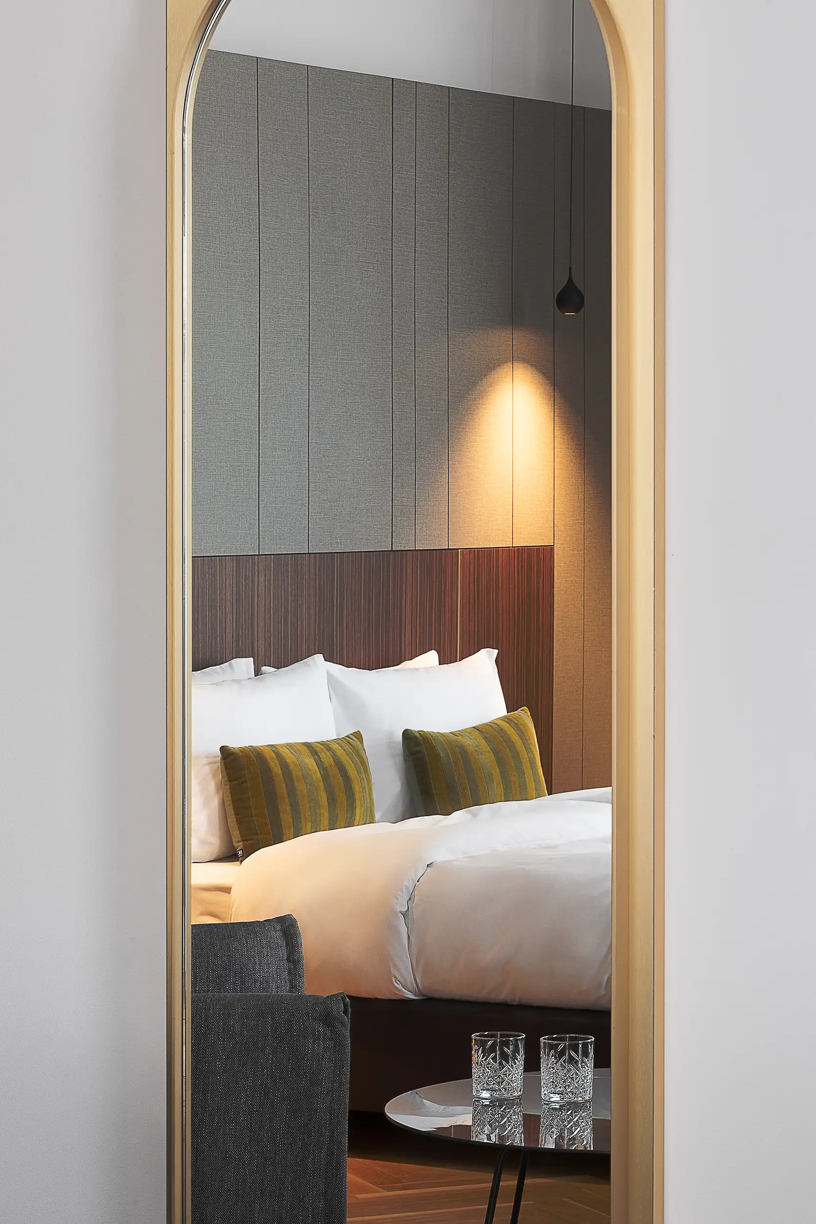 A luxurious king-size bed and a small table with cut glasses are visible through a minimalist, golden-bordered mirror in the Deluxe Comfort Suite at Numa Laurel in Prague, Czech Republic.