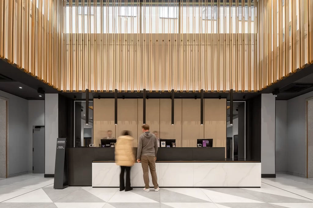 Die Frontalansicht der minimalistischen Rezeption des Radisson Blu Prag mit hohen Decken, goldenen Wanddekorationen und verschwommenen Gästen davor.