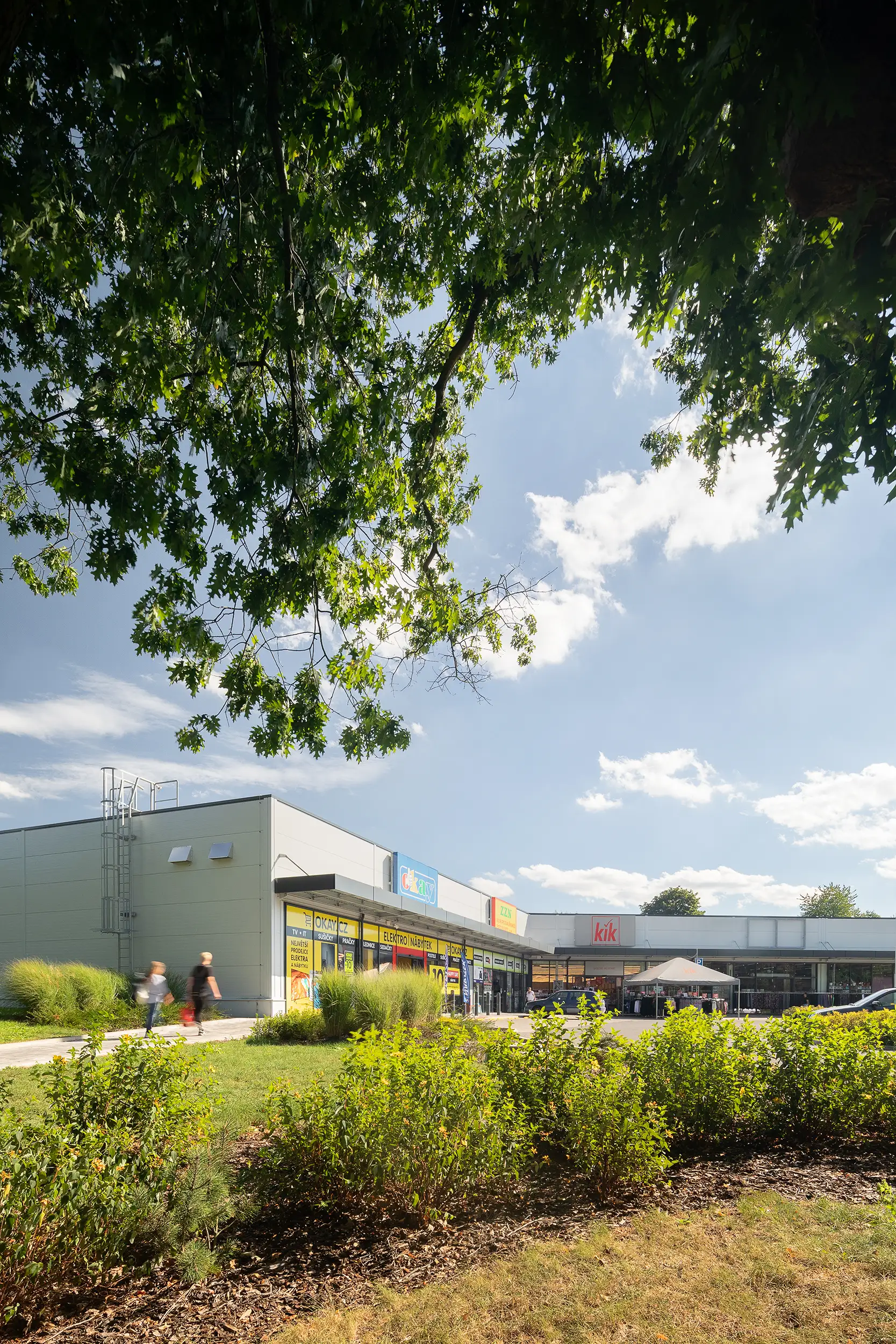 The daylight vertical photo of the Retail Park in Choceň captured through the greenery in the foreground with blurred people approaching the stores of Okay and KiK.