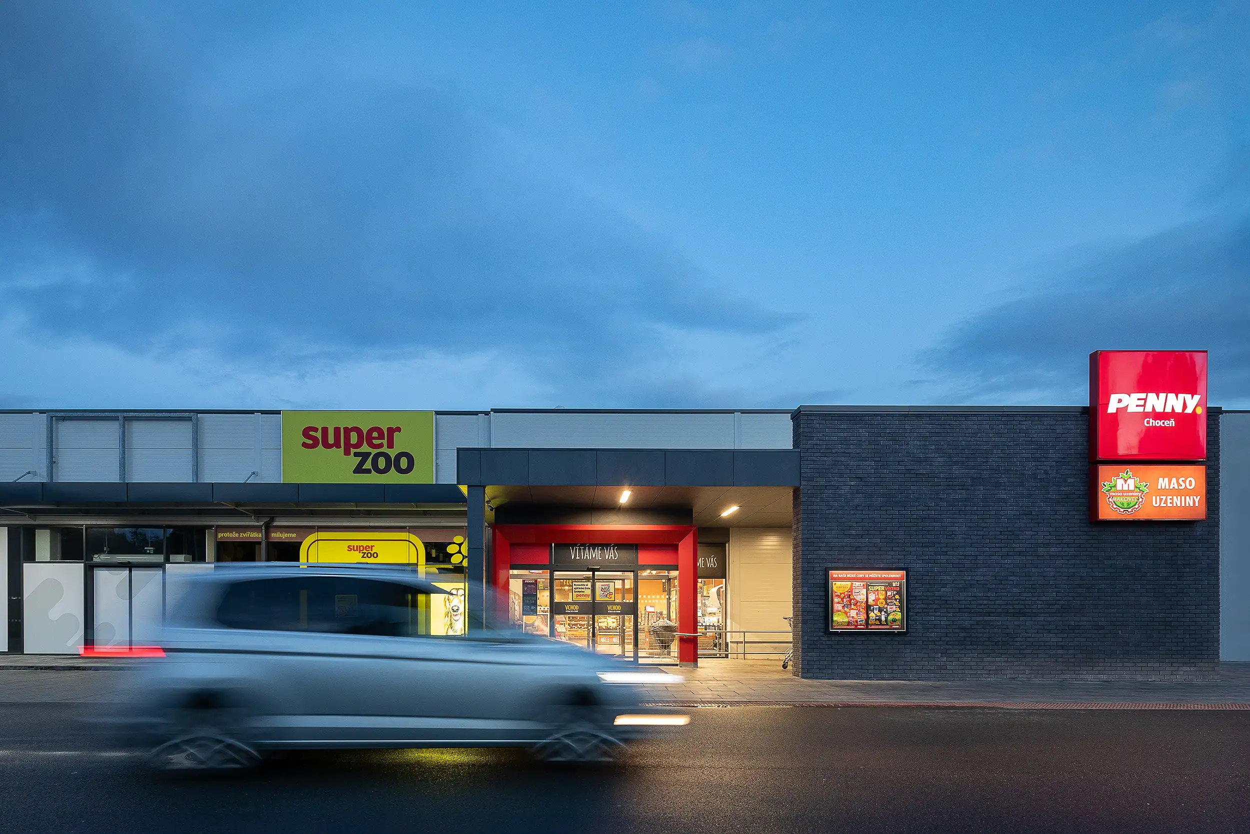 Image of the entrance to Penny Market captured during the blue hour with a blurred car passing by in the foreground, located in commercial space in Choceň.