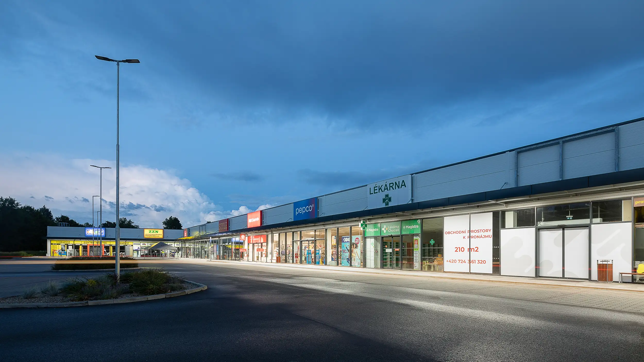 The overlook of the illuminated shopping area in Choceň, Czech Republic, captured during dusk, shows the stores, empty parking lot, and public lights off.