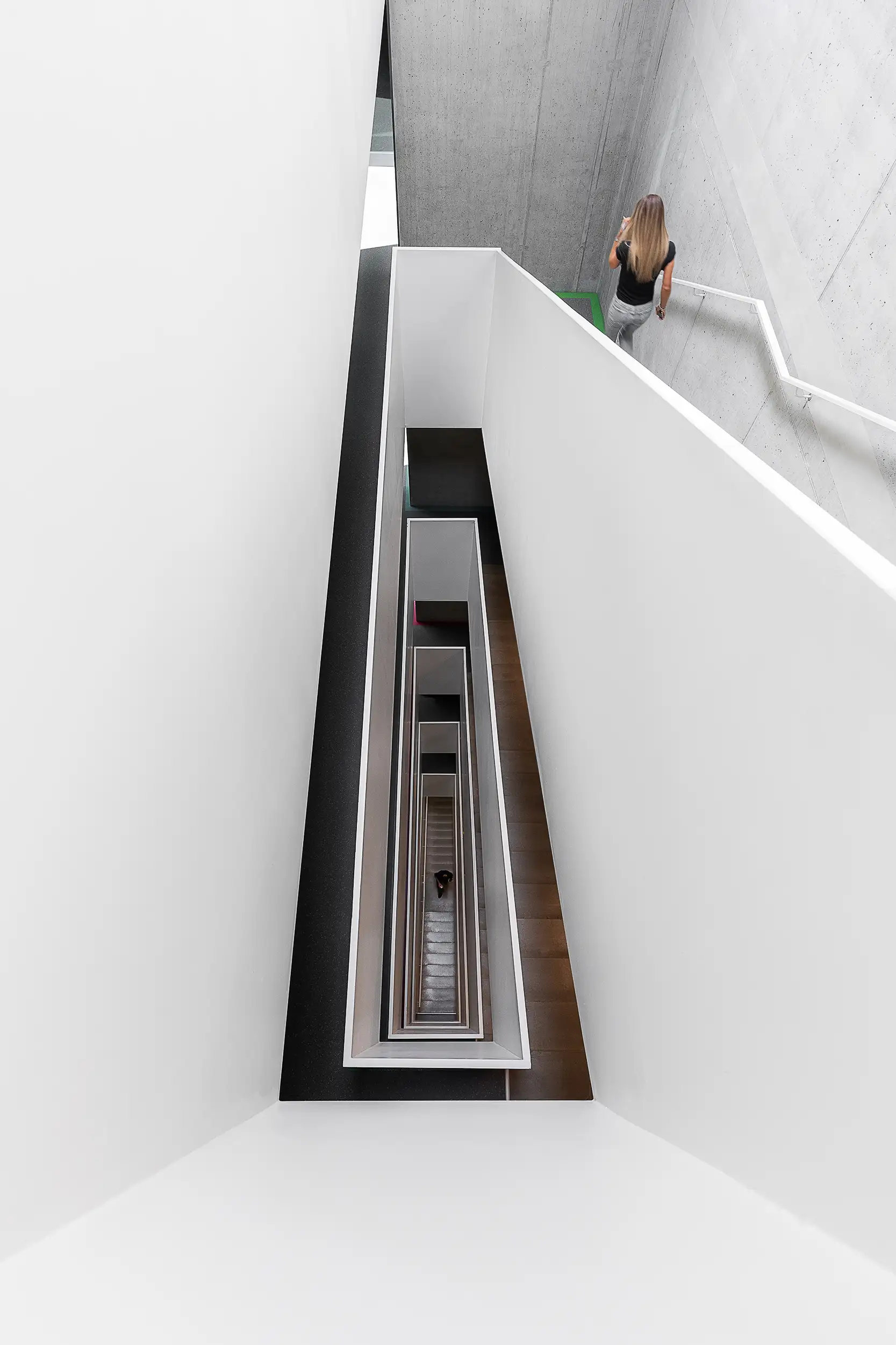A top-view photograph of a rectangular interior spiral staircase in a contrasting black and white color scheme combined with visual concrete also showcases a woman blurred in motion descending downwards.