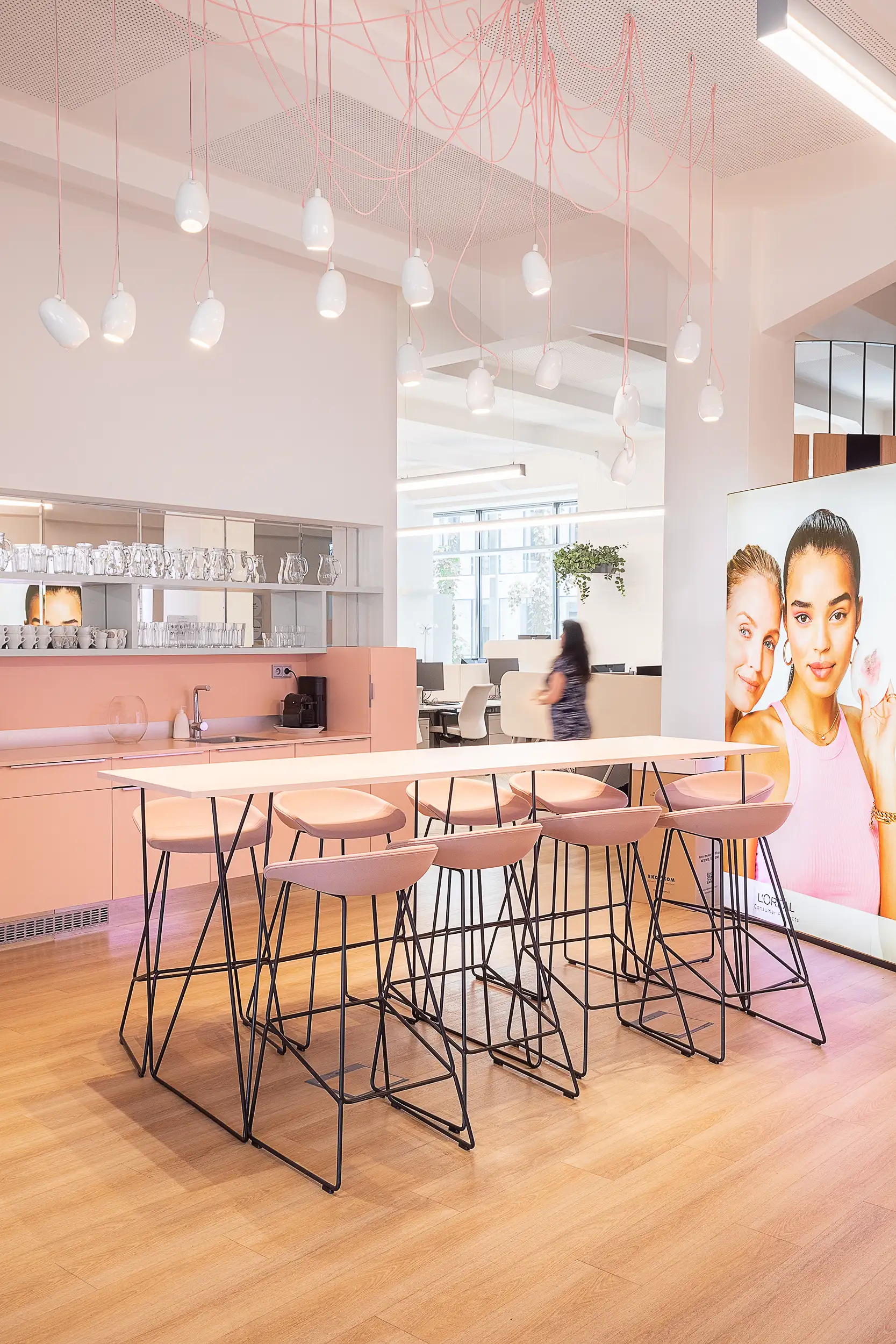 A pink-tinted office kitchen with a table and chairs on thin black legs is illuminated by designed lights hanging from tangled cables at L'Oréal in Palace Křižík located in Prague, Czech Republic.