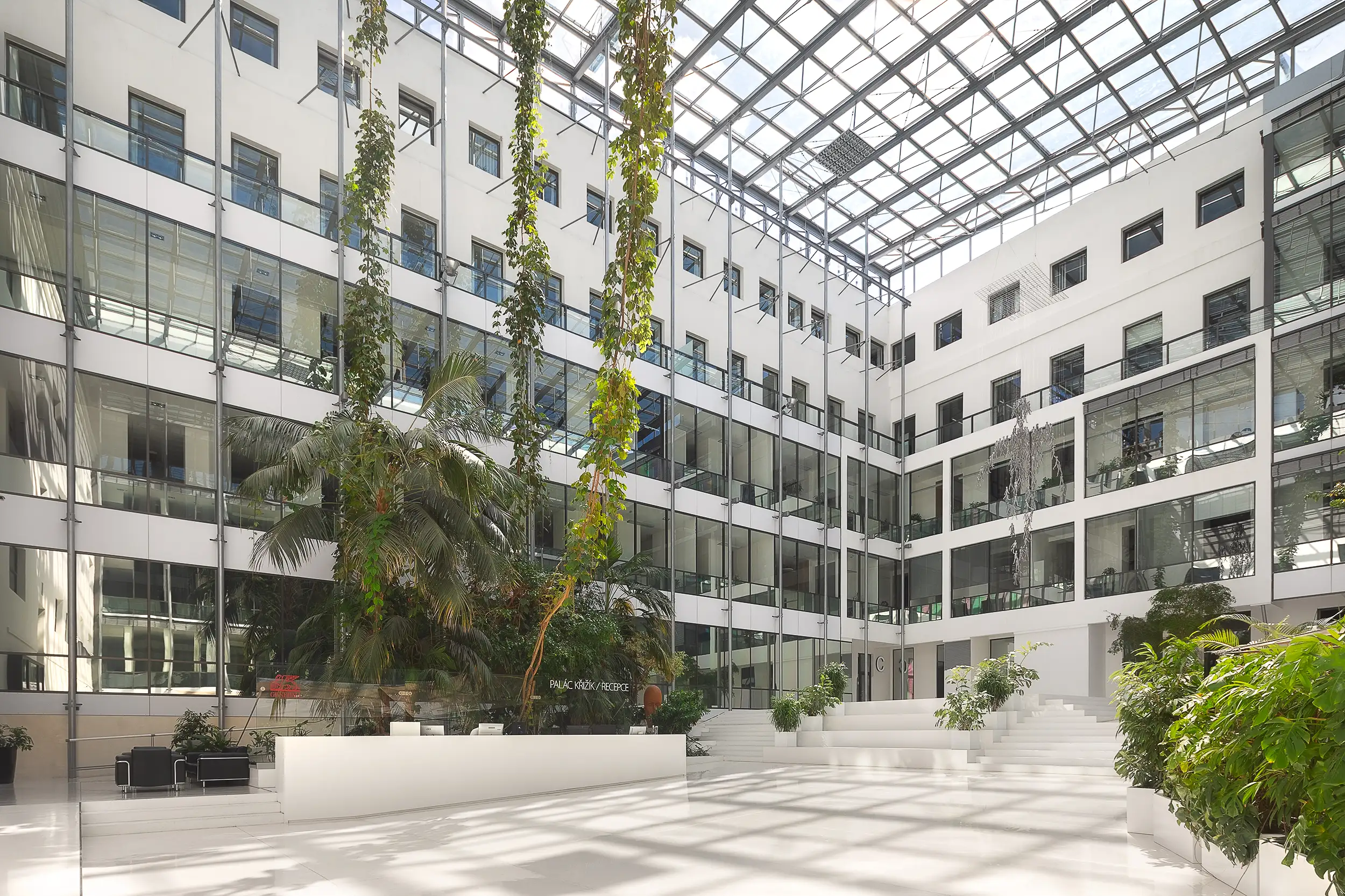 Here is a photograph of the spacious, minimalist, modern atrium inside the Křižík Palace in Prague, Czech Republic. The interior, created by AHK Architects, is entirely white and adorned with plants hanging from the ceiling.