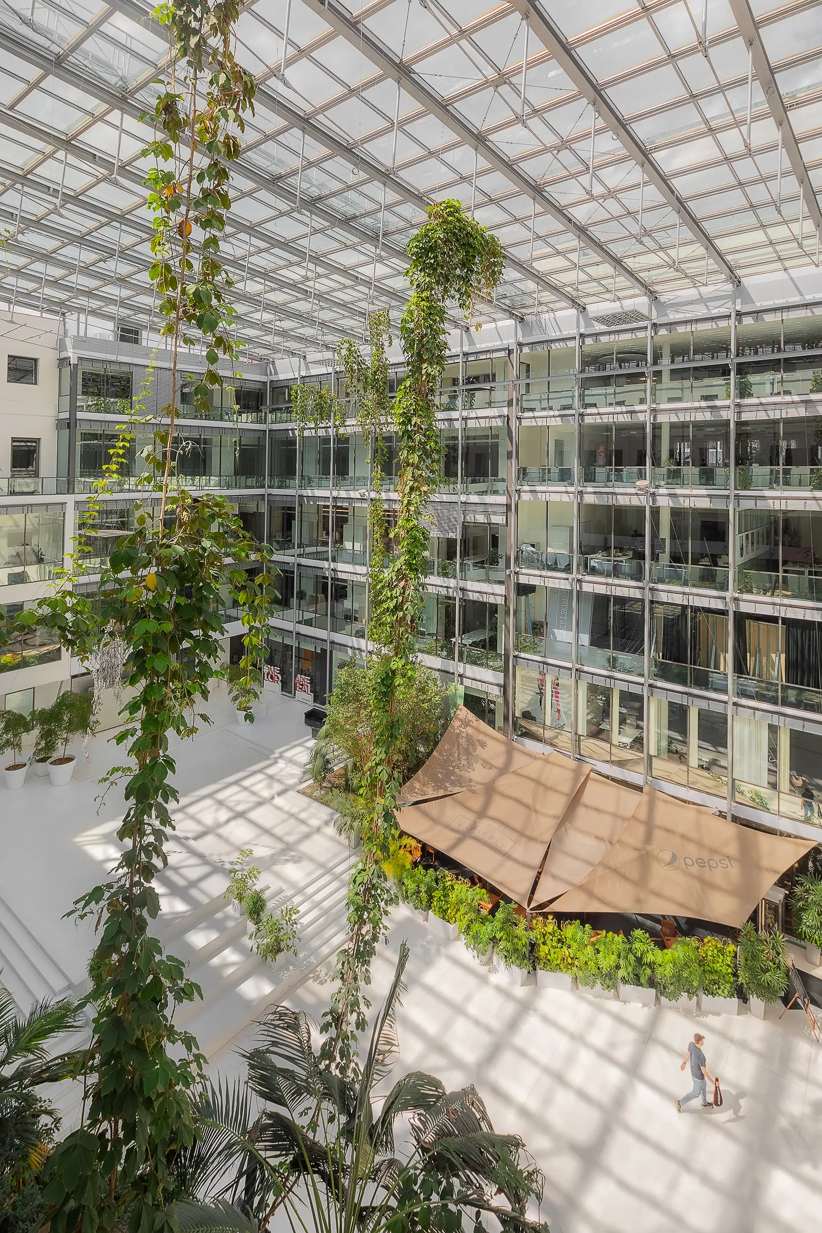 A portrait oriented photograph of the white interior atrium with a glass roof at Křižík Palace in Anděl, Prague, Czech Republic shows its height and work with hanging greenery. The atrium was designed by AHK Architects.