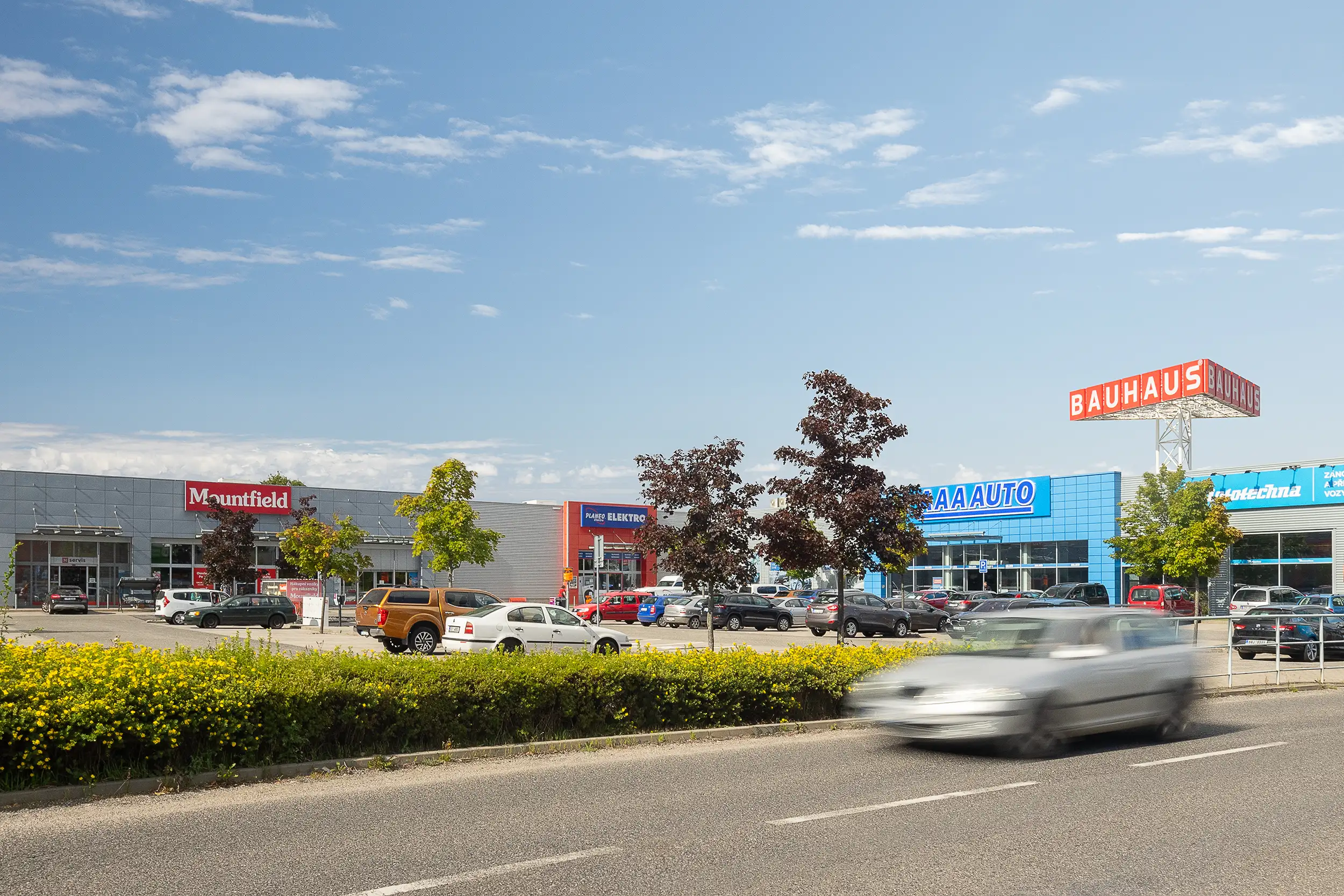 A daylight overview photo of Retail Park Severka in Liberec captured across the street with cars passing by, blurred in motion, containing the Mountfield, Planeo Elektro, and AAA auto stores.