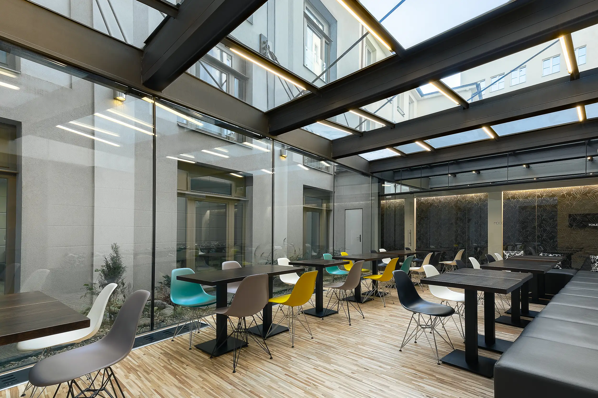 Wintergarten im Hotel MOODs Boutique Prag mit Glasdach, filigraner Stahlkonstruktion und modernem Sitzbereich mit farbigen Designstühlen.
