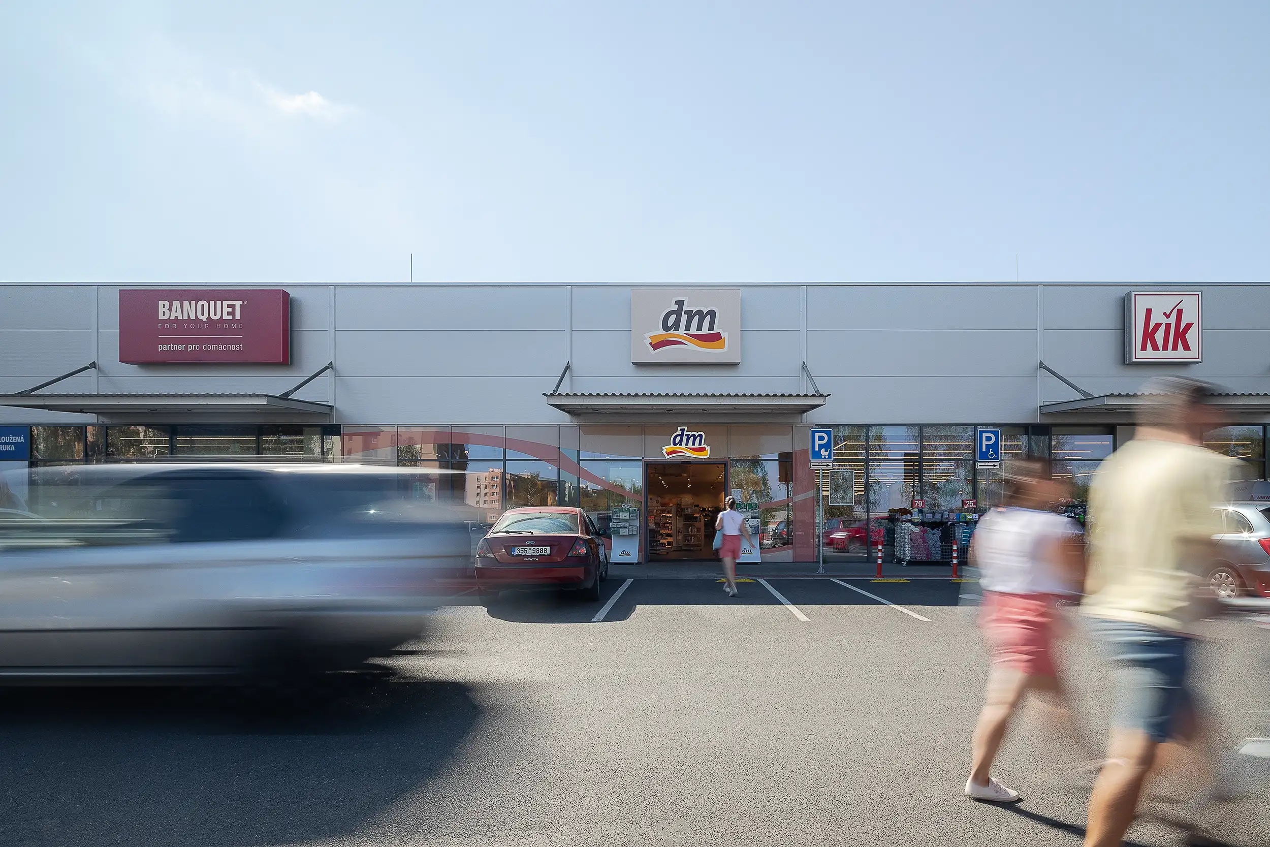 Photography of the retail center in Milevsko focuses on the entrance to the DM store, framed by moving people and cars in motion from the left and right.