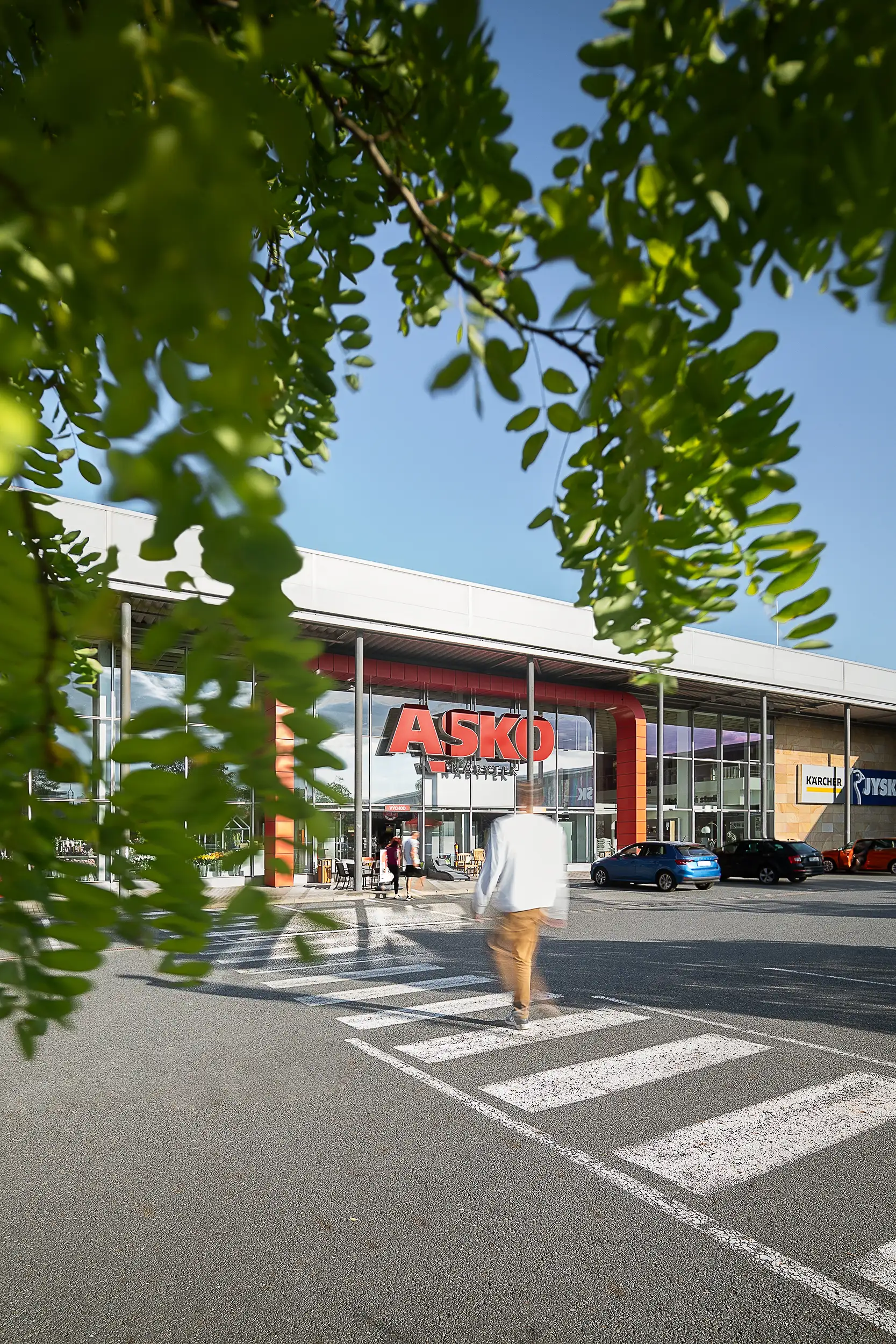 A man, blurred by the motion in the picture, is walking toward the ASKO furniture store entrance in the commercial area of Mladá Boleslav, Czech Republic near a pedestrian crossing. The greenery frames the top of the image.