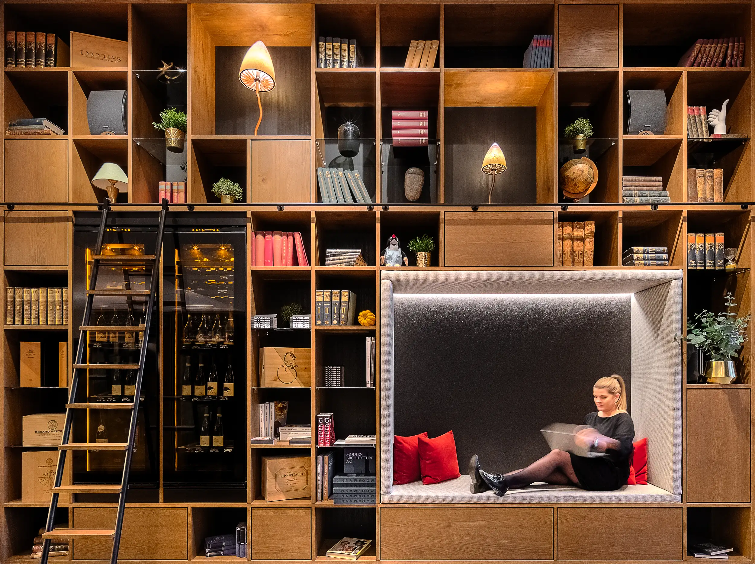 A frontal photo of the bookshelf wall in the lobby of Mosaic House in Prague, Czech Republic, consists of artistic lights in the form of mushrooms, flowers, books, and a blurred woman working at her laptop.