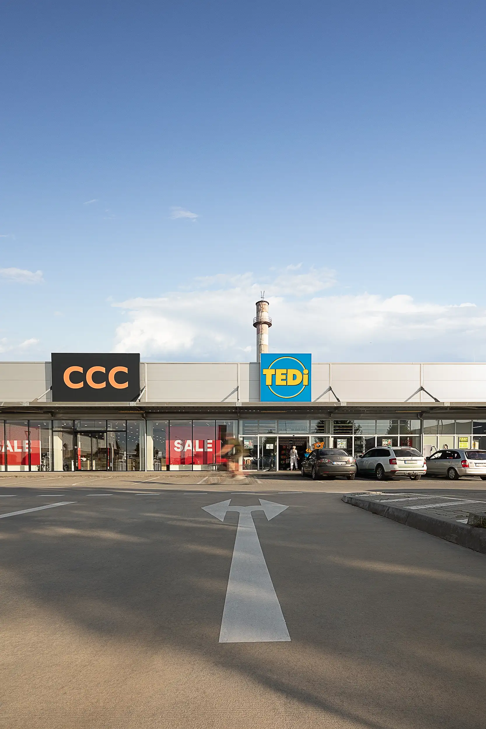 Photo of a retail park in Senica, Slovakia, captured from a lower perspective, containing traffic arrows on the road pointing to a blurry cyclist riding in front of CCC and TEDi stores.
