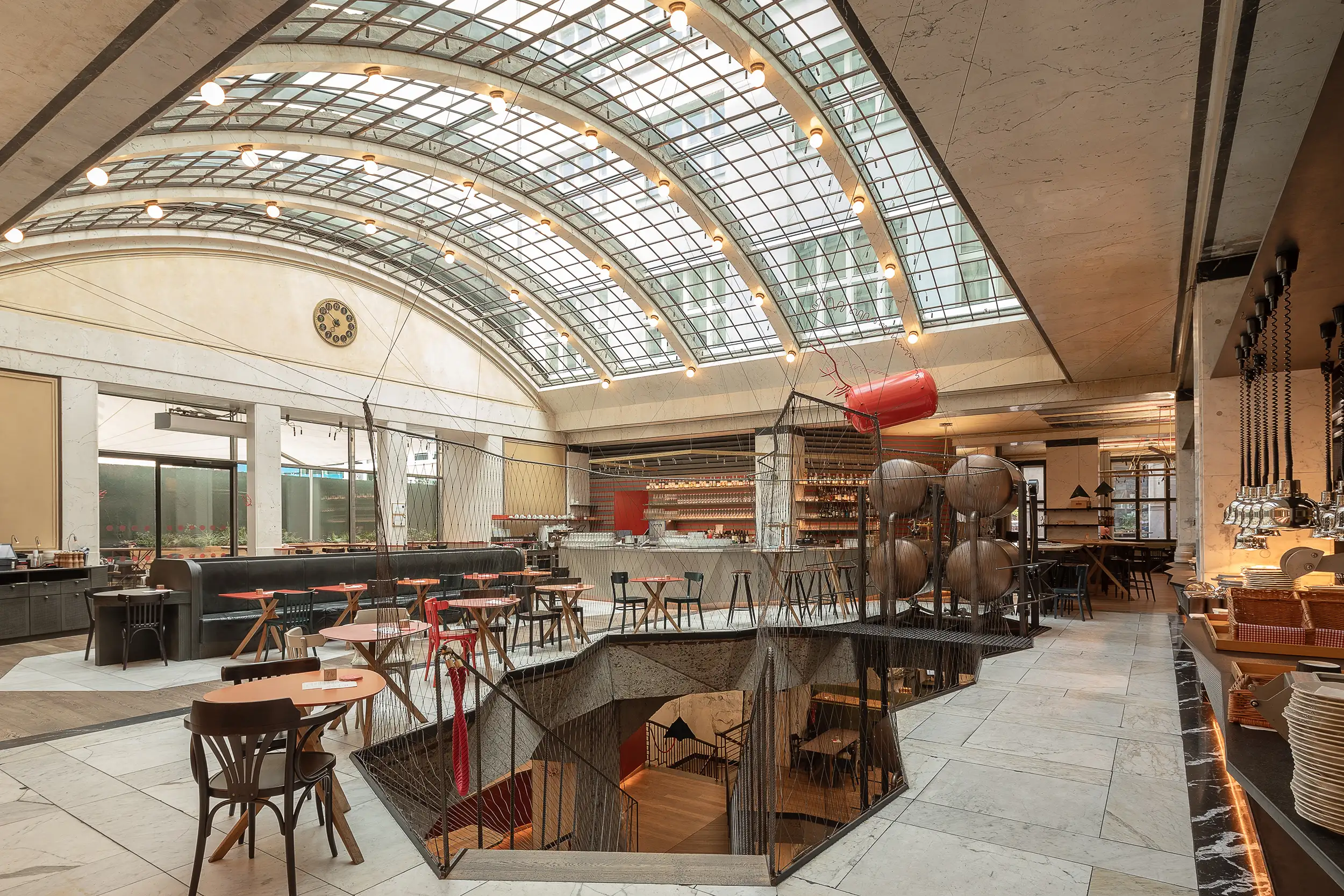 Wide interior photography of restaurant Červený Jelen in Prague, Czech Republic, situated in the hall of the former bank with an arched glass roof and staircase to the lower floors, where the vaults used to be.