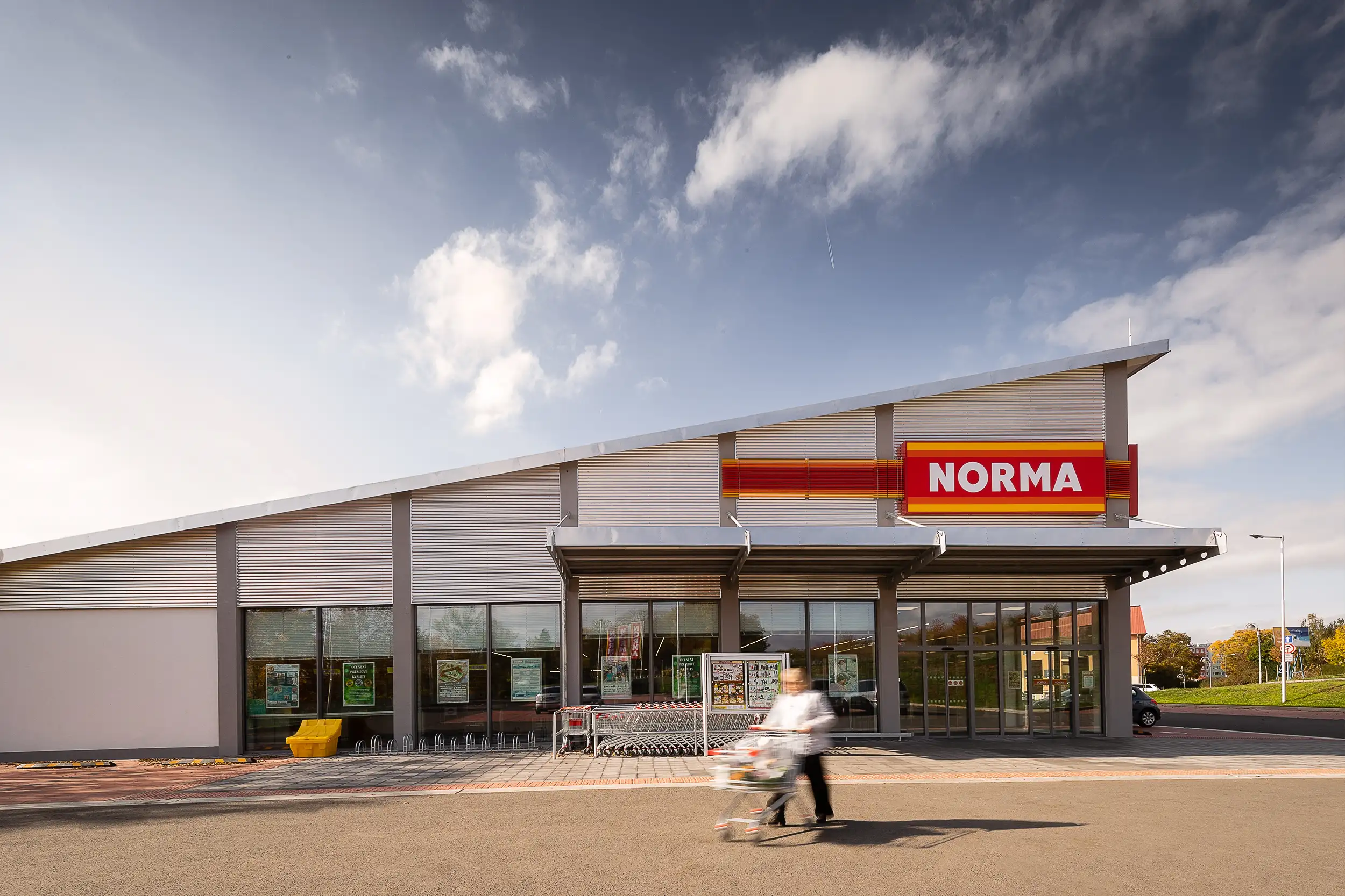 Hero photo of the Norma shopping store in Pobořany, Czech Republic, with a pensioner with a shopping trolley blurred in motion in the foreground.