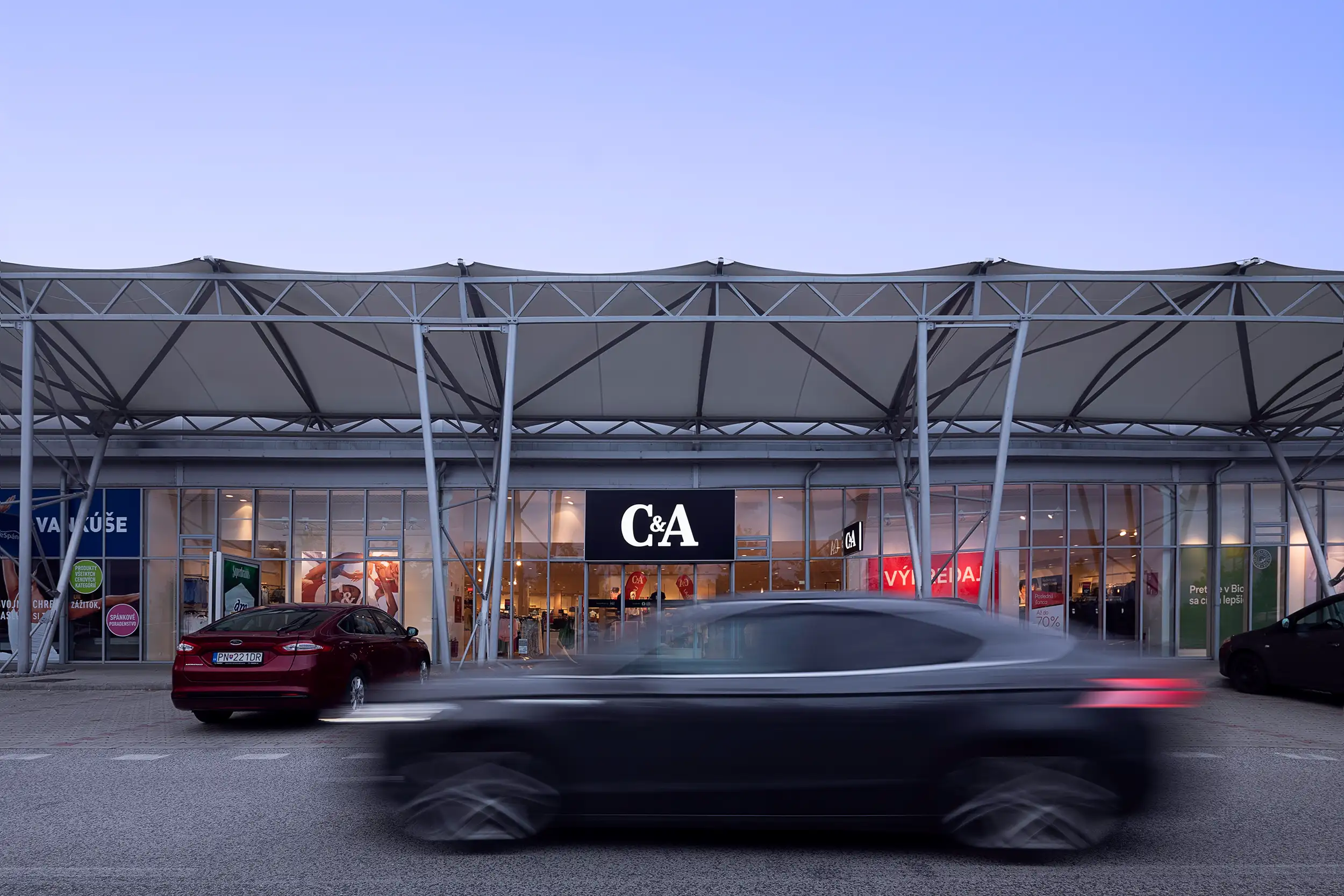 A dusk shot of an entrance to an H&M store in the MALL Trnava, Slovakia, captures the building with a passing car blurred by the motion in the foreground.