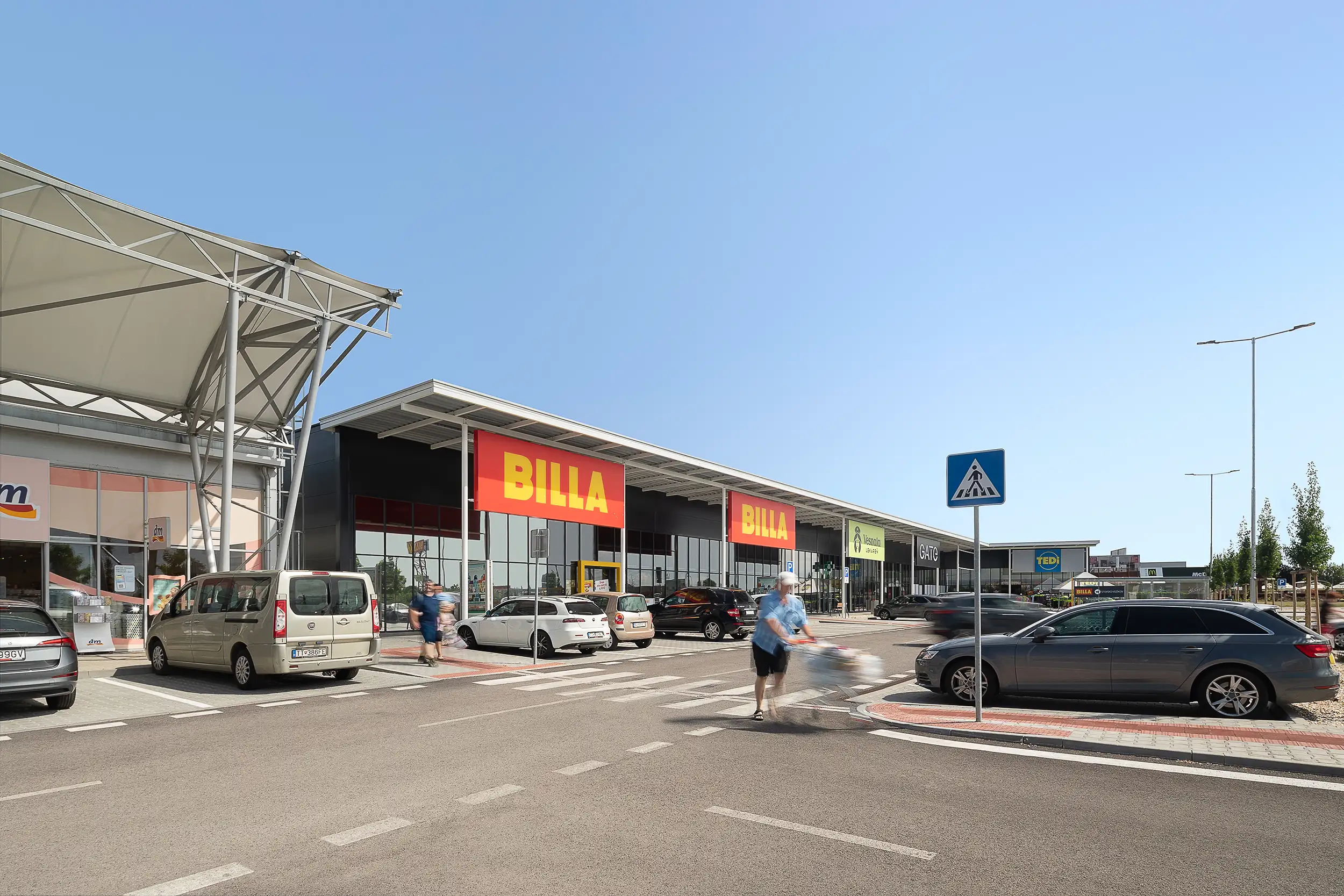 Daylight photography of the freshly built second part of the retail park in Trnava, Slovakia, capturing mainly the BILLA store and people blurred in motion.