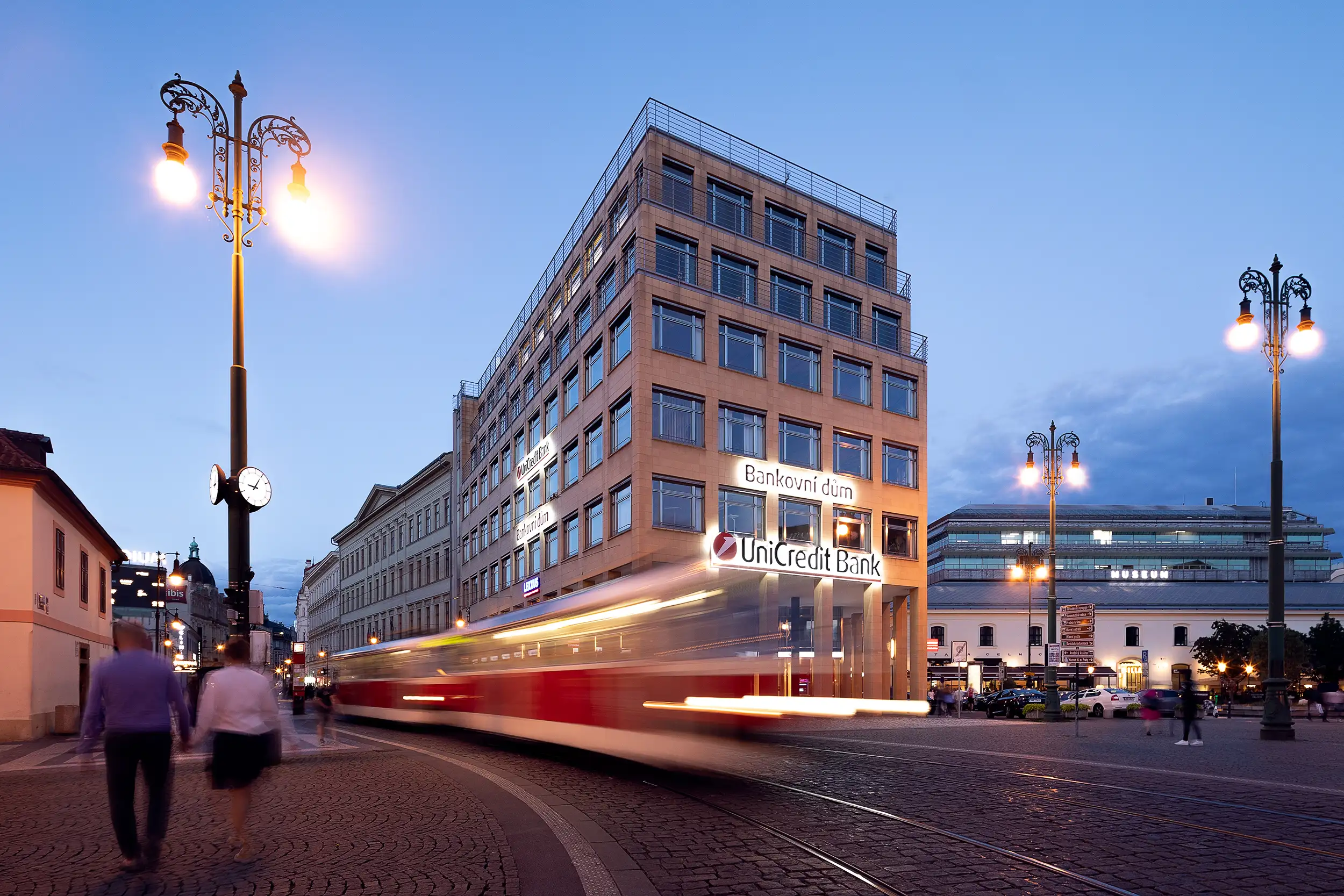 During the blue hour, a long-exposure photo was taken from Na Porici Street showing a tram and people moving in front of UniCredit Bank's Banking House on Republic Square in Prague.