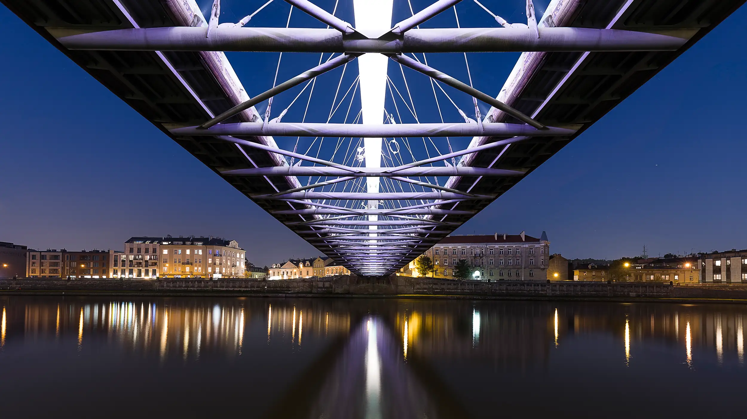 A perfectly symmetrical night photo taken from beneath Father Bernatek’s Bridge in Krakow displays the illuminated bridge with its mirror image on the Visla River surface alongside the riverfront on the opposite site.