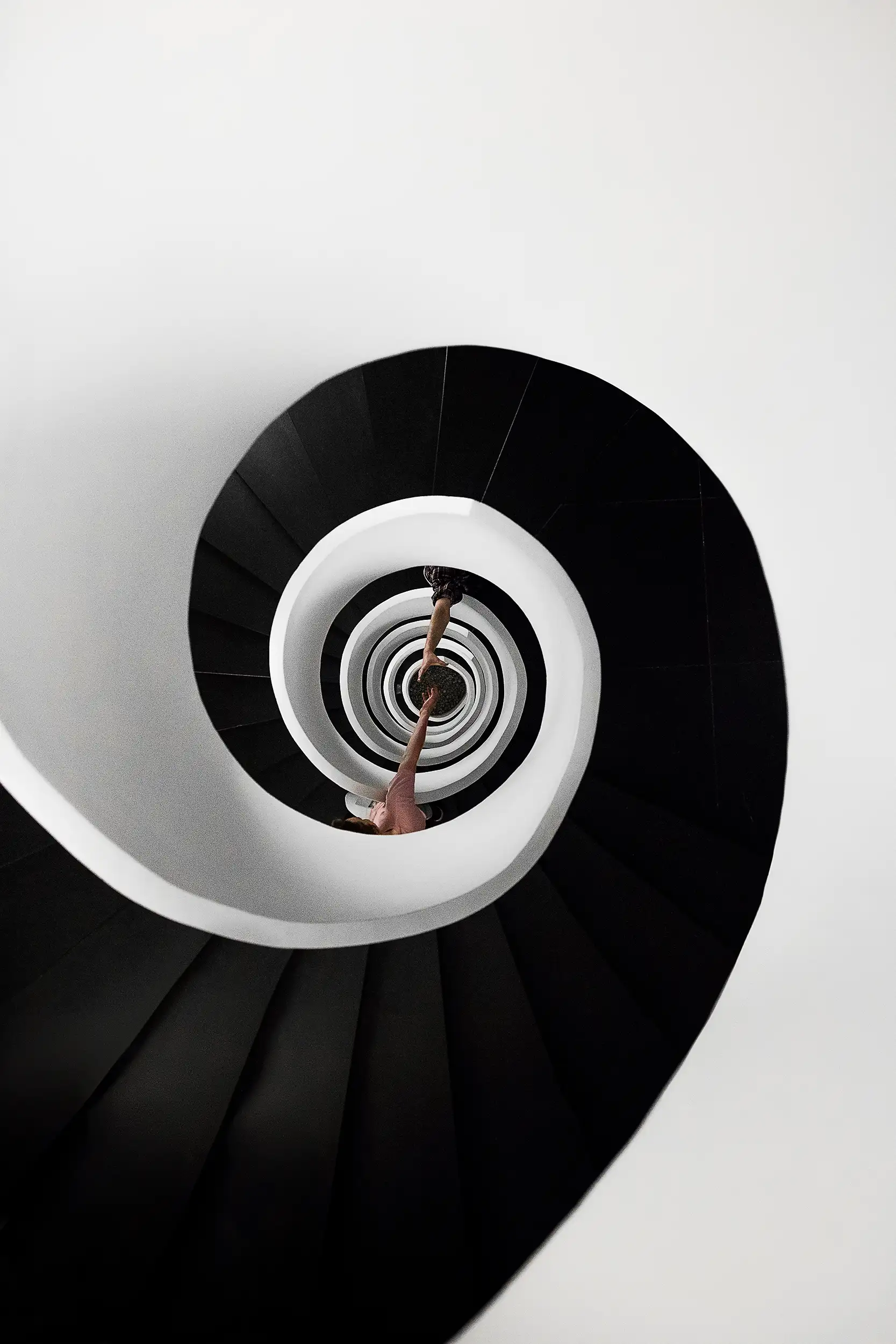 Fine art architecture photography captures the downward view of two people trying to hold their hands across the spiral staircase.