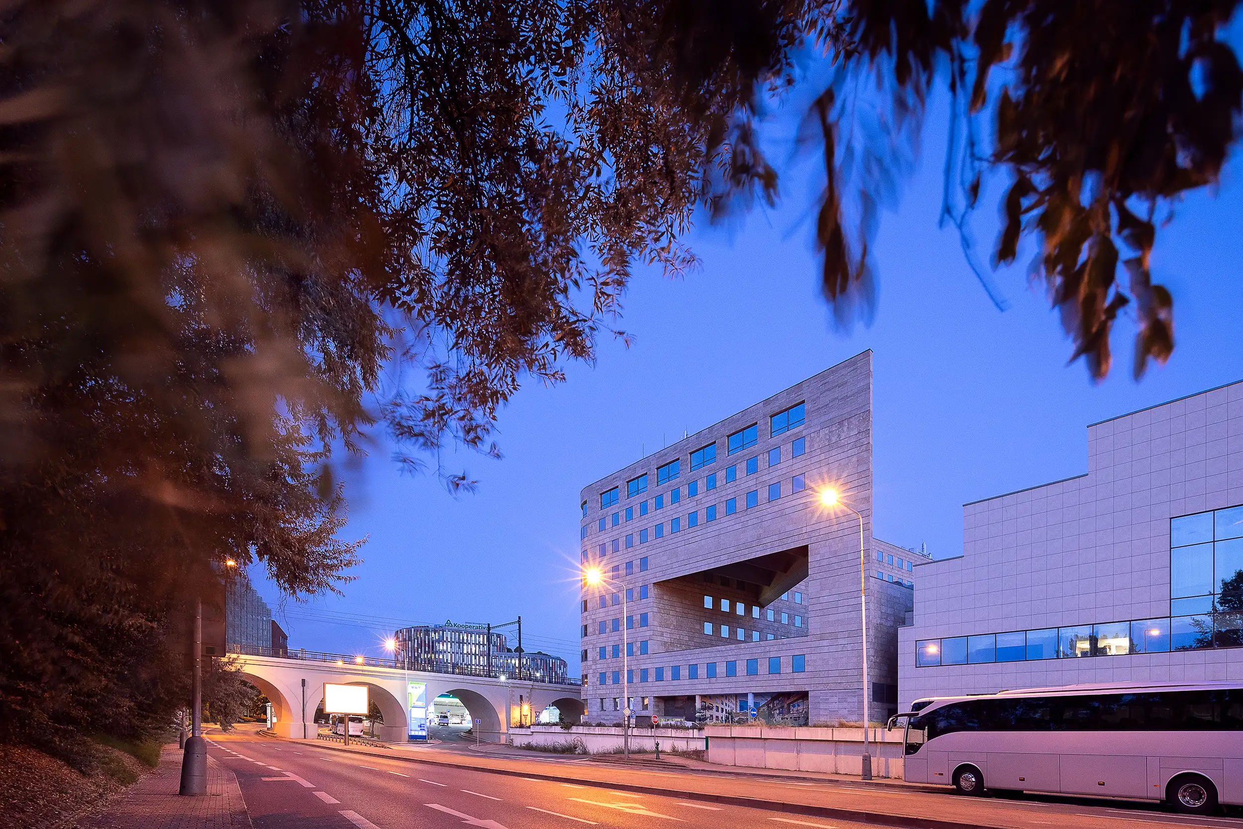 Gesamtansicht des International Business Centre Prag bei Dämmerung im Kontext des Karlíner Negrelli-Viadukts mit bewegungsverschwommenen Blättern im Vordergrund.