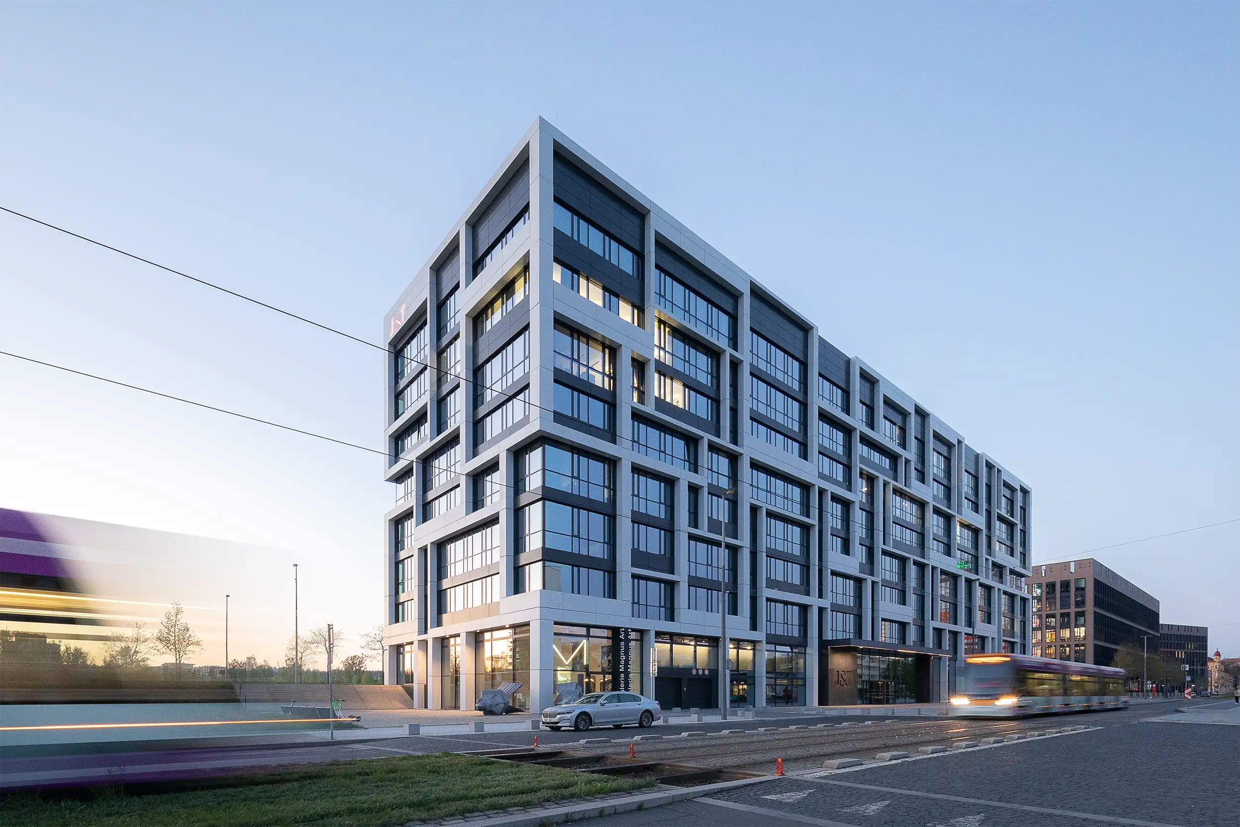 Gesamtansicht des neuen Prager Hauptsitzes der J&T Bank von der Sokolovská-Straße bei Dämmerung mit vorbeifahrenden Straßenbahnen im Vordergrund.