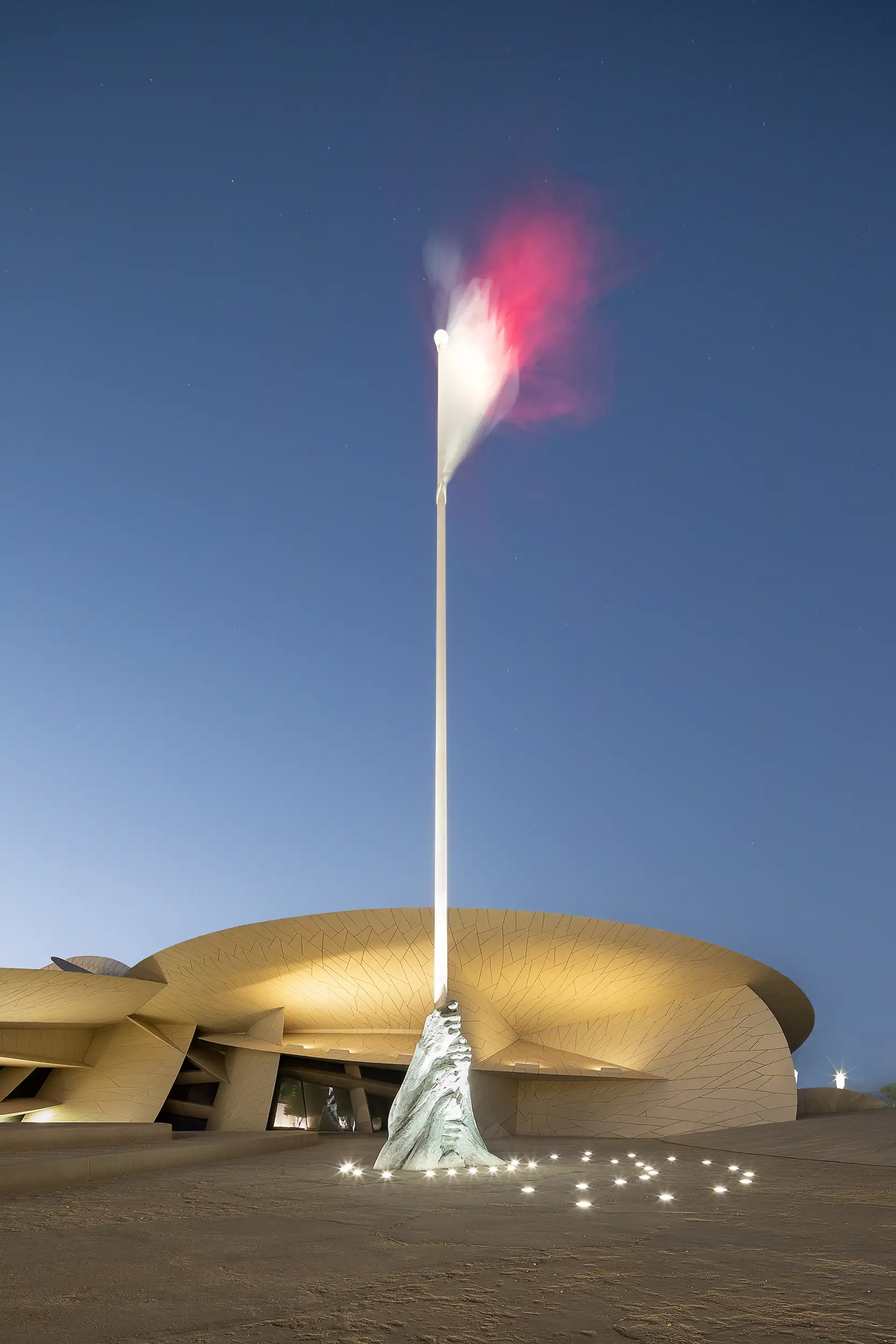 A flagpole with a motion blurred Qatar flag with the Doha National Museum building in the background captured at night.