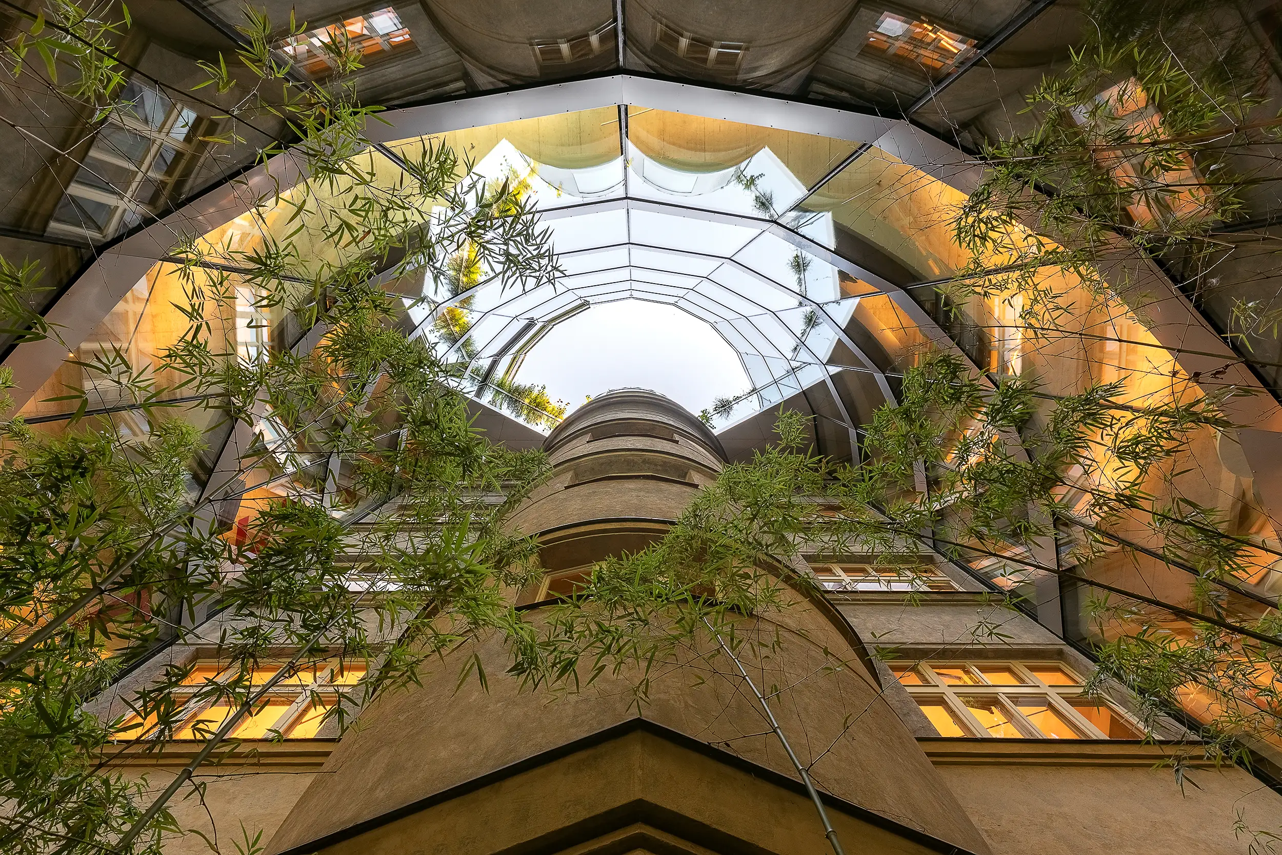 Upward view of the inner courtyard of the Špork Palace in Prague, featuring a glass roof and bamboo plants growing in the courtyard.