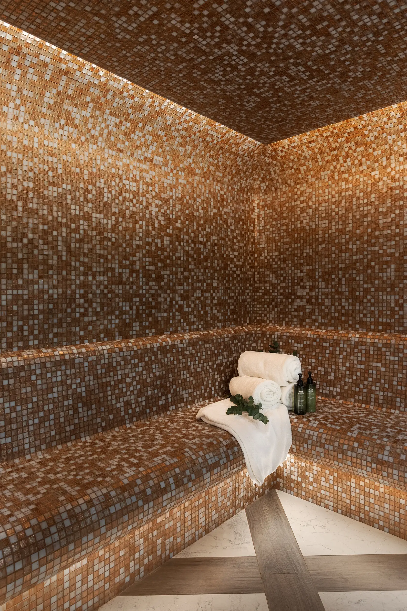 Vertical shot of the steam sauna, lined with small mosaic tiles, complete with towels, shower gels and body lotions.