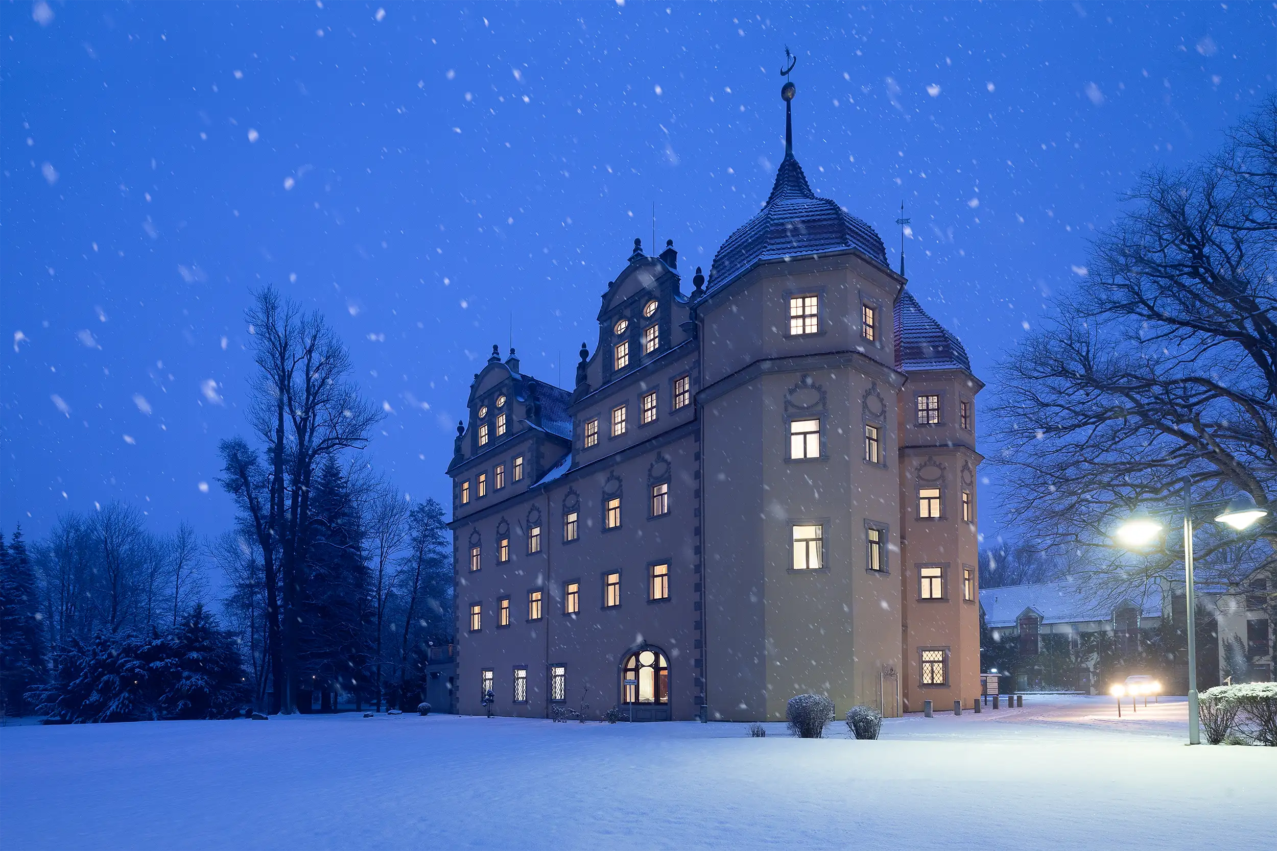 Side view of illuminated Schloßhotel Althörnitz during the night, surrounded by a serene blanket of snow during snowfall.
