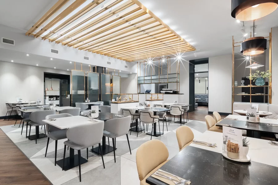 Elegantní a minimalistická hotelová restaurace v Radisson Blu Praha laděná do světlých barev s bílými prvky, doplněná výraznými zlatými doplňky a stropními dekoracemi.