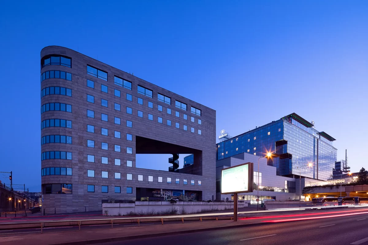 Gesamtansicht des International Business Centre (IBC) Prag bei Dämmerung im Kontext des Rohanské-Ufers und des Hilton Hotels mit Lichtspuren des Straßenverkehrs.