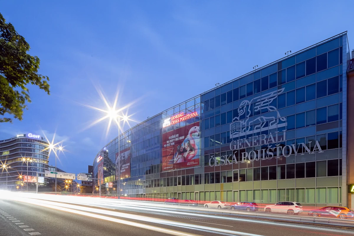 Noční fotografie hlavní fasády komplexu Pankrác West, pražské centrály Generali, obalené souvislou krycí stěnou s nápisy a tónovanými skly, zachycená z ulice 5. května, doplněná o světelné stopy projíždějících aut