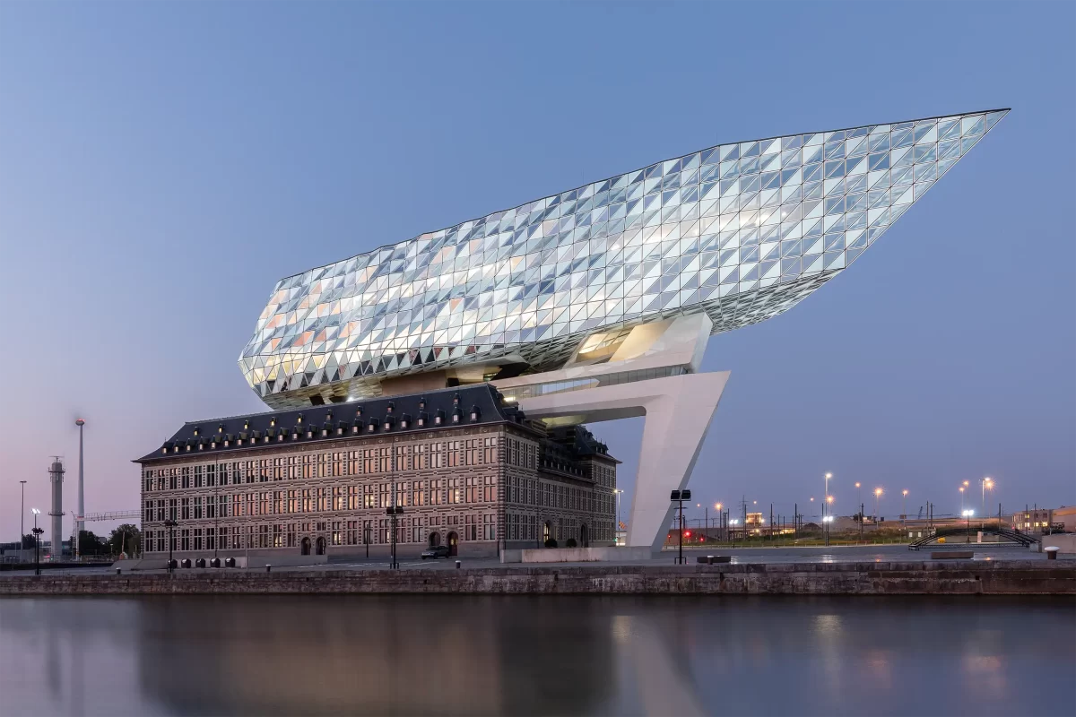 Gesamtansicht des Port House Antwerpen von Zaha Hadid bei Dämmerung mit historischem Gebäude und futuristischem Glasanbau.