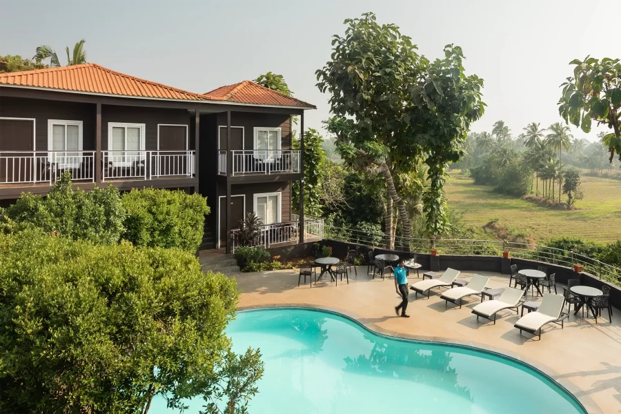 Bazénová část resortu s lehátky, obklopená bujnou zelení a dvoupatrovou budovou s balkony s výhledem na tropická pole.“