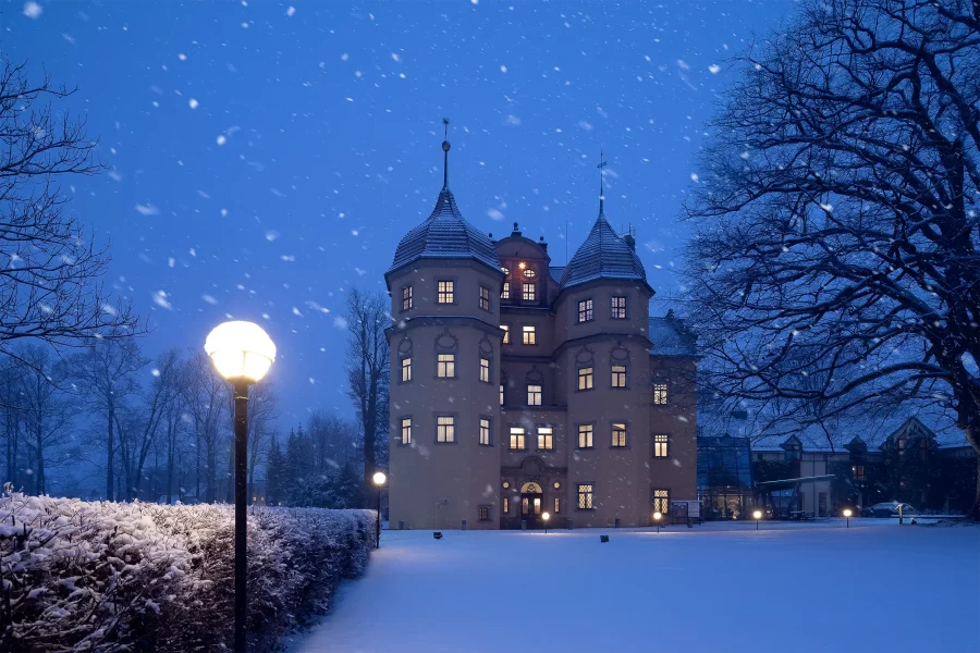 Beleuchtetes Schlosshotel Althörnitz bei Nacht mit Schneefall und historischer Parklampe im Vordergrund.